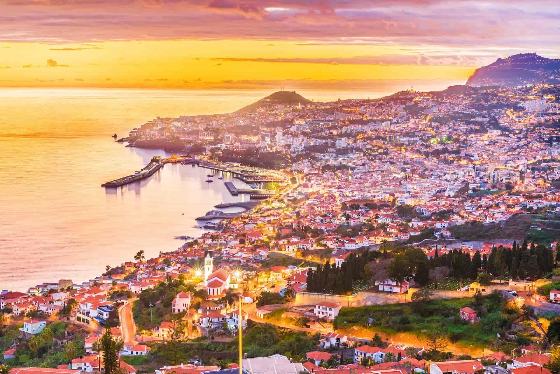 Funchal city lights along the coast at sunset with warm orange and purple sky over the sea.
