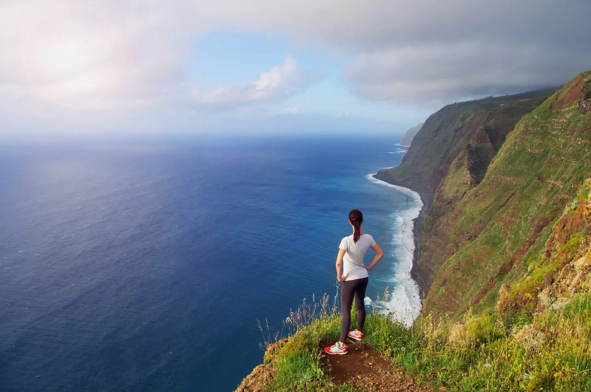 Woman hiking overlooks vast blue Atlantic Ocean from steep, green coastal cliffs.