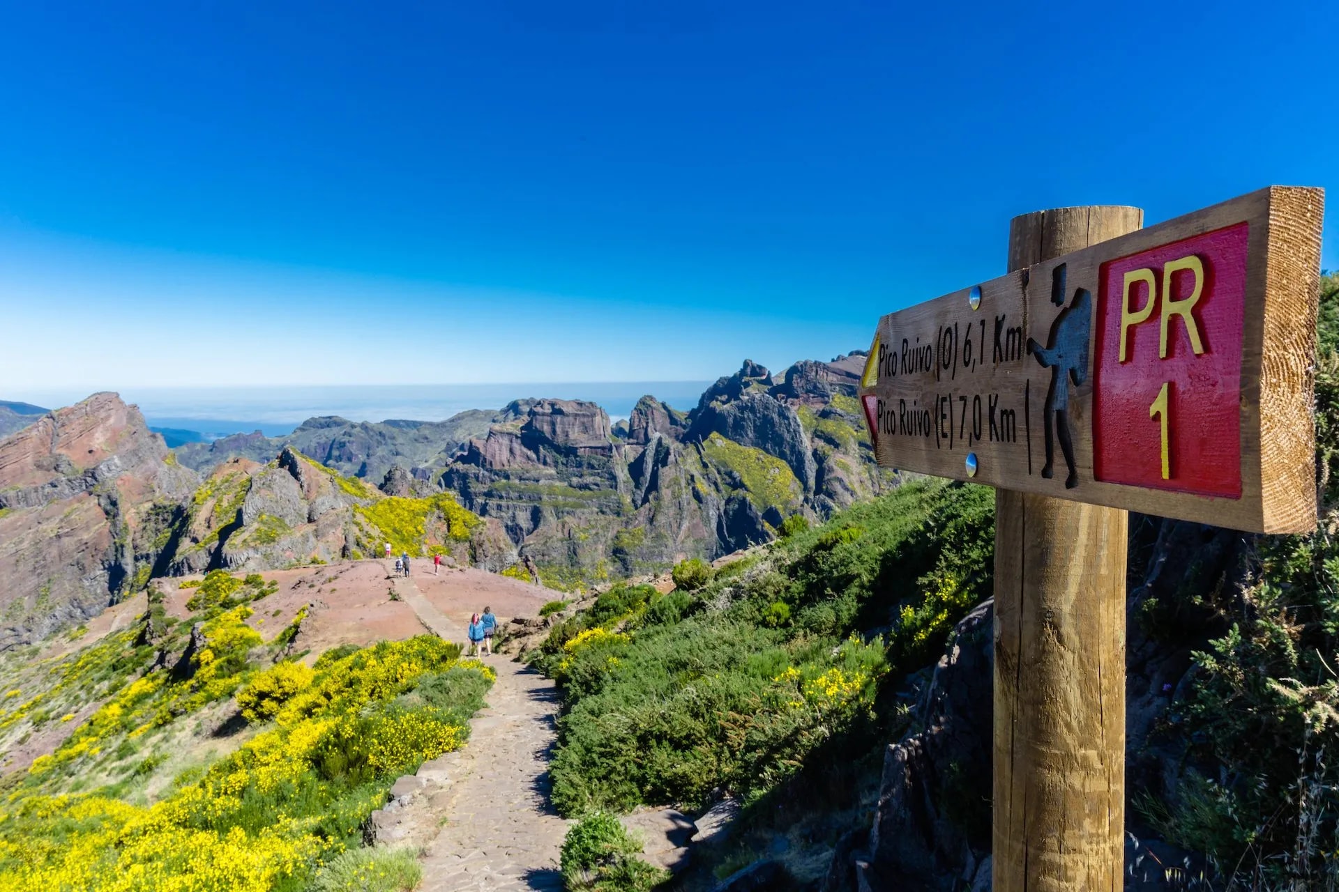 Hiking trail sign for PR 1 Pico Ruivo with hikers on path overlooking rugged mountains.