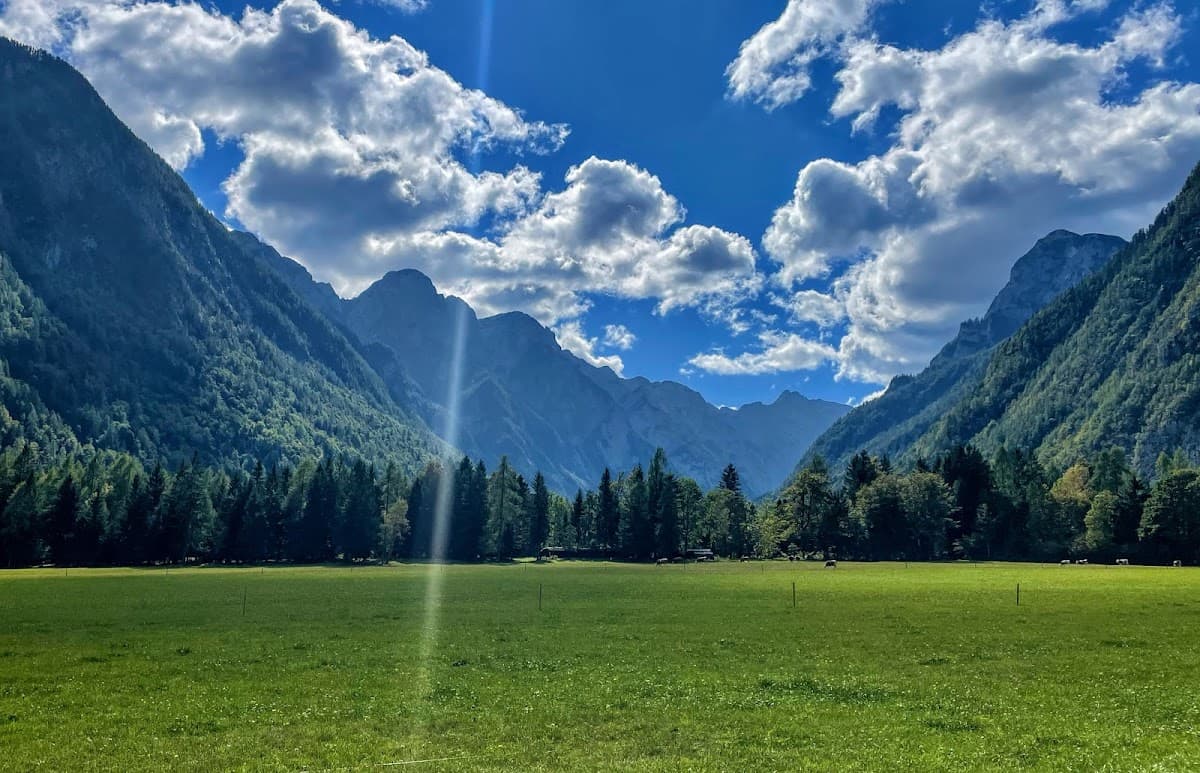 Green meadow valley floor framed by steep, forested mountains under a bright blue sky with clouds.