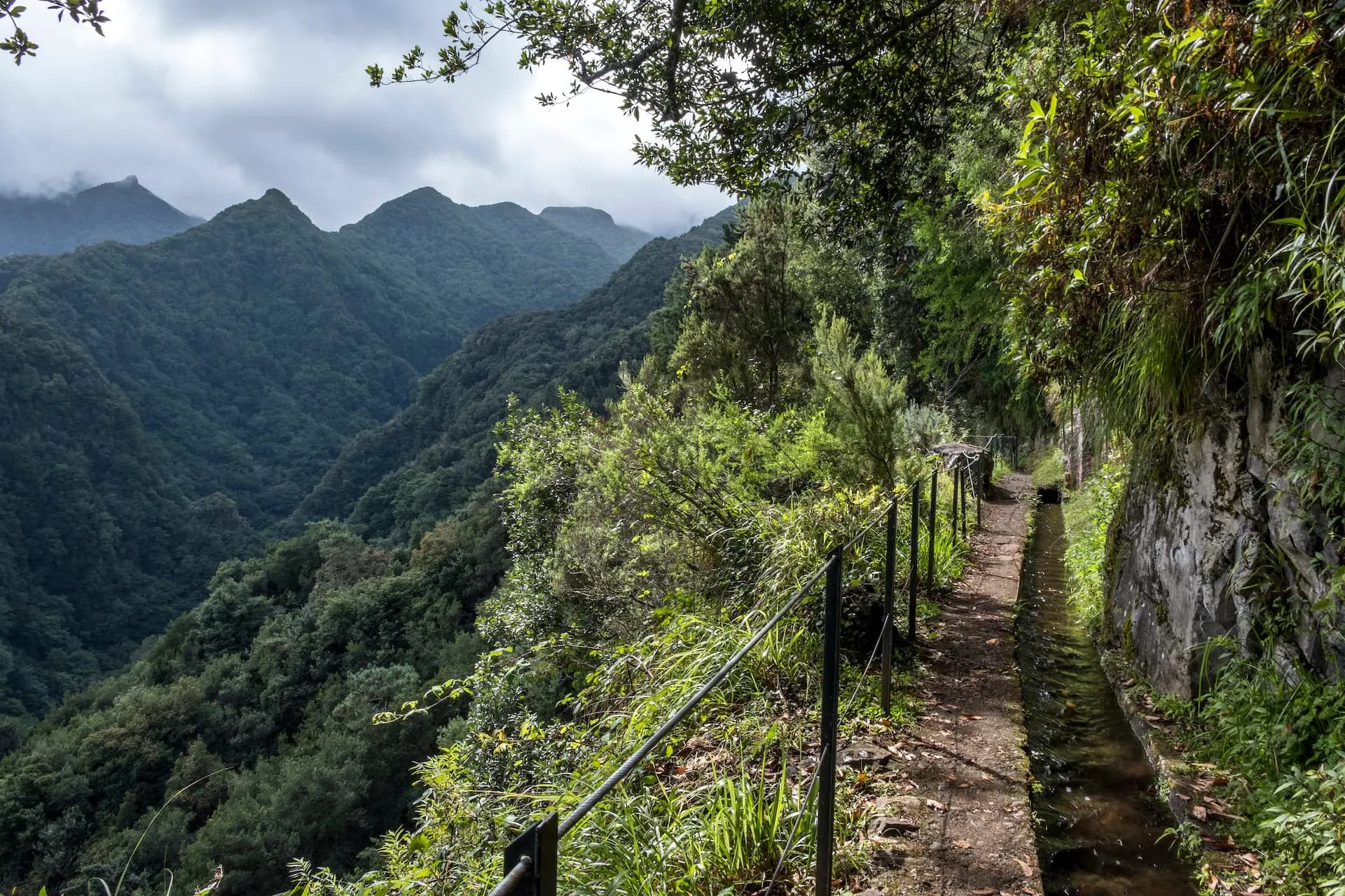 Hiking path alongside a levada irrigation channel overlooking lush, forested mountains under cloudy skies at Levada do Rei.