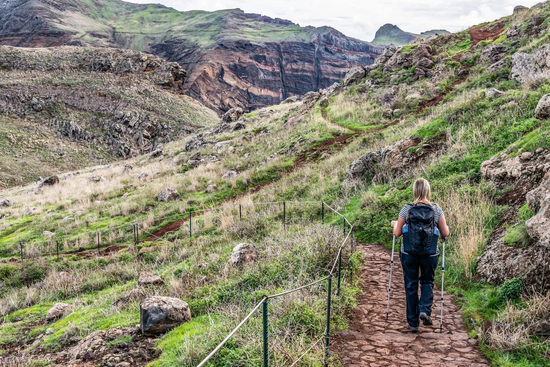 Hiking on Ponta de São Lourenço trail with woman using poles past rocky, grassy slopes.