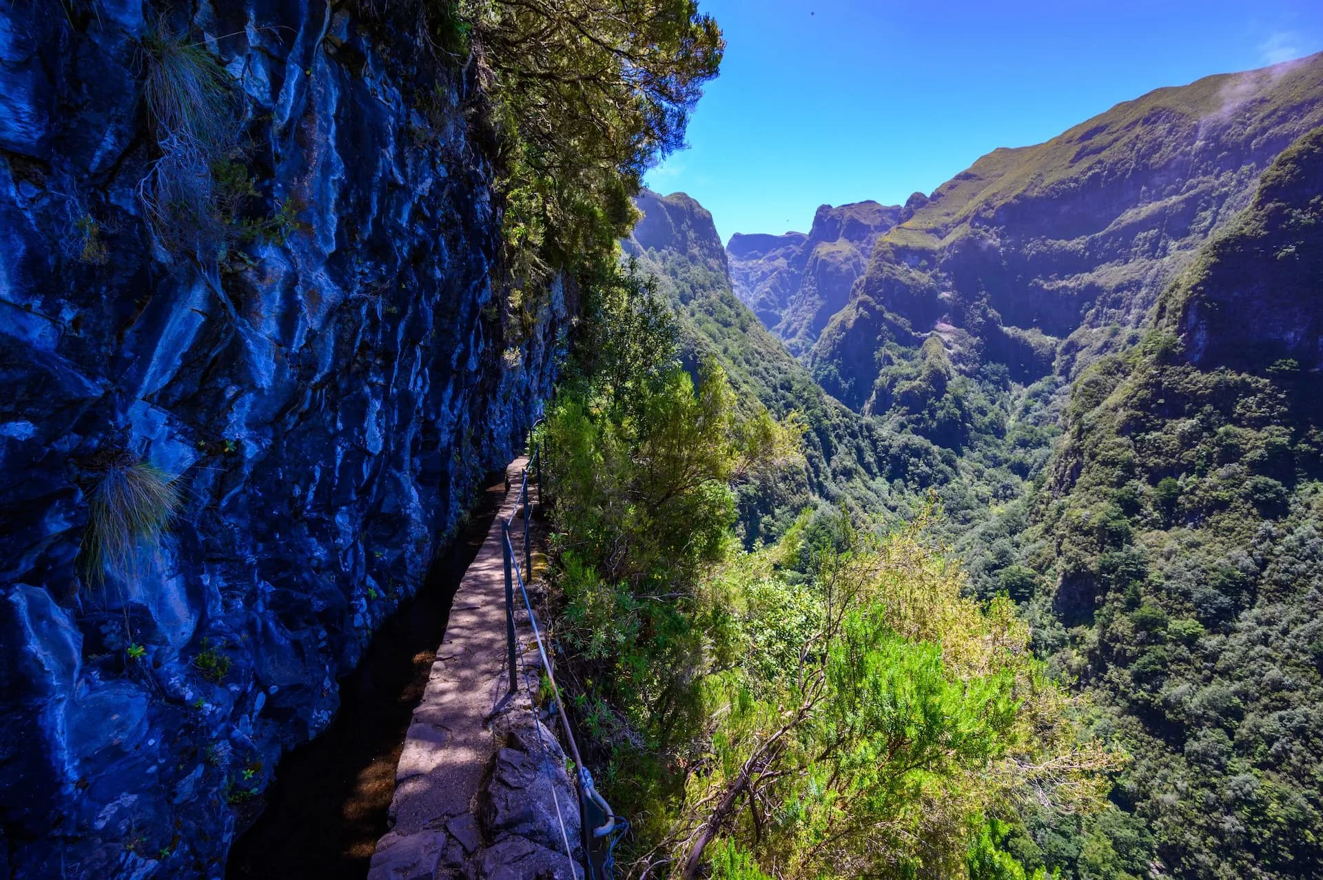 Hiking along the Levada do Caldeirão path next to a dark blue rock face overlooking a lush green valley.