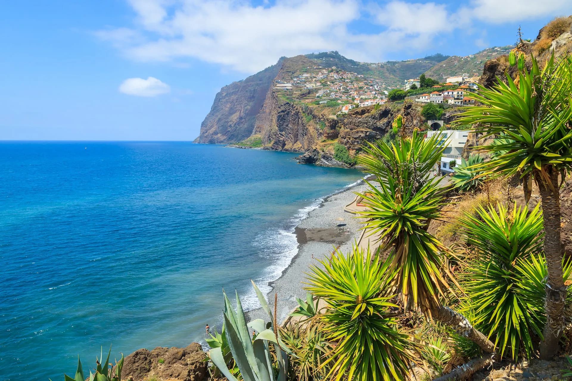 Coastal view of Cabo Girao with steep cliffs, pebble beach, blue ocean, and town on hillside.