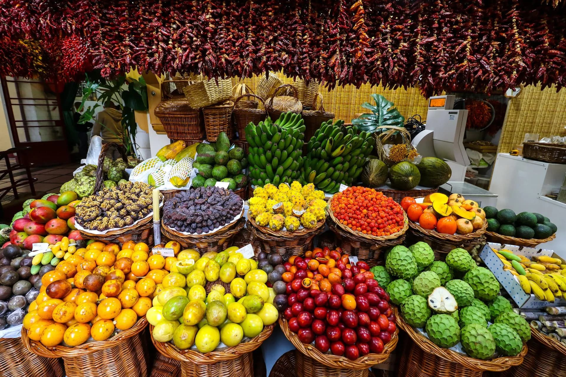 Abundant fresh fruit display under hanging dried red peppers at Mercado dos Lavradores.